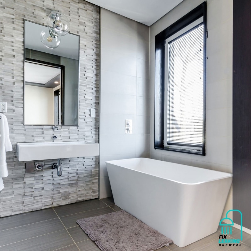 Modern bathroom with freestanding white tub, gray tile walls, large mirror, and natural light—perfect for leaking shower repair.